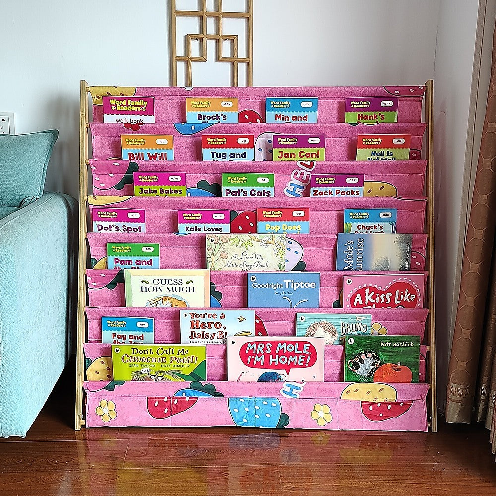 Pink fabric children's book display rack filled with picture books in room setting next to teal sofa and decorative screen