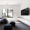 Fixed TV wall bracket lifestyle in minimal white room with grey sofa black window and two round ottomans
