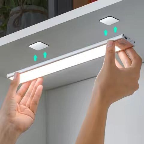 Two hands attaching LED bar light to underside of cabinet using magnetic mounts shown with green upward arrows
