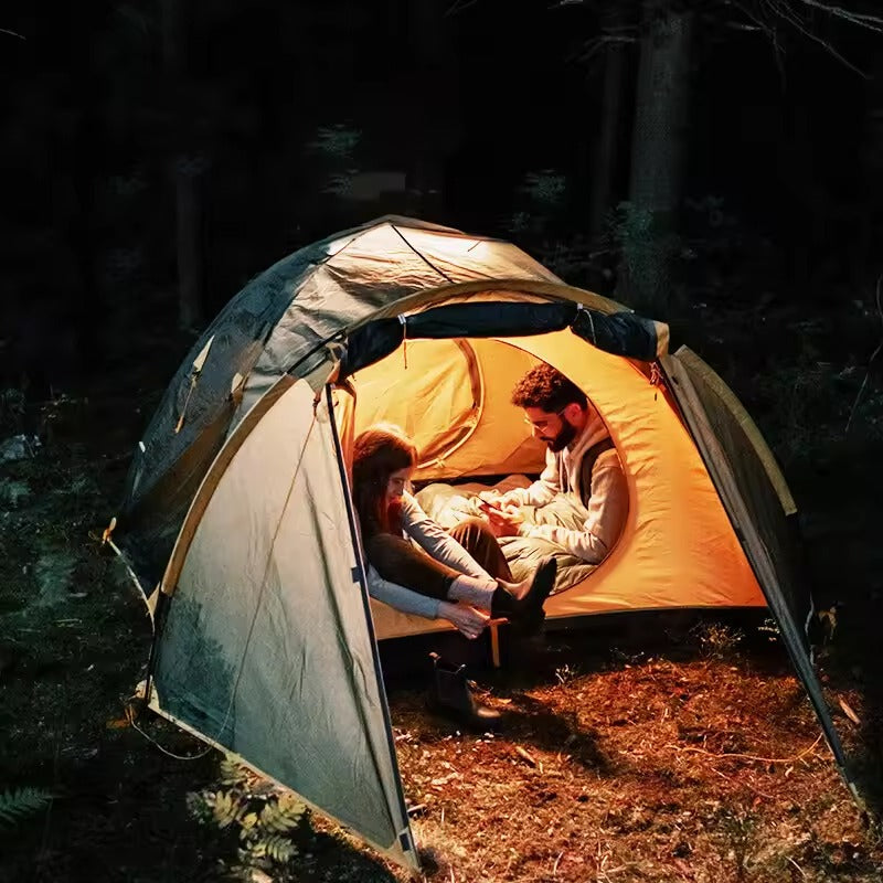 Two people inside illuminated dome tent at night in dark forest, warm orange glow from camping light inside
