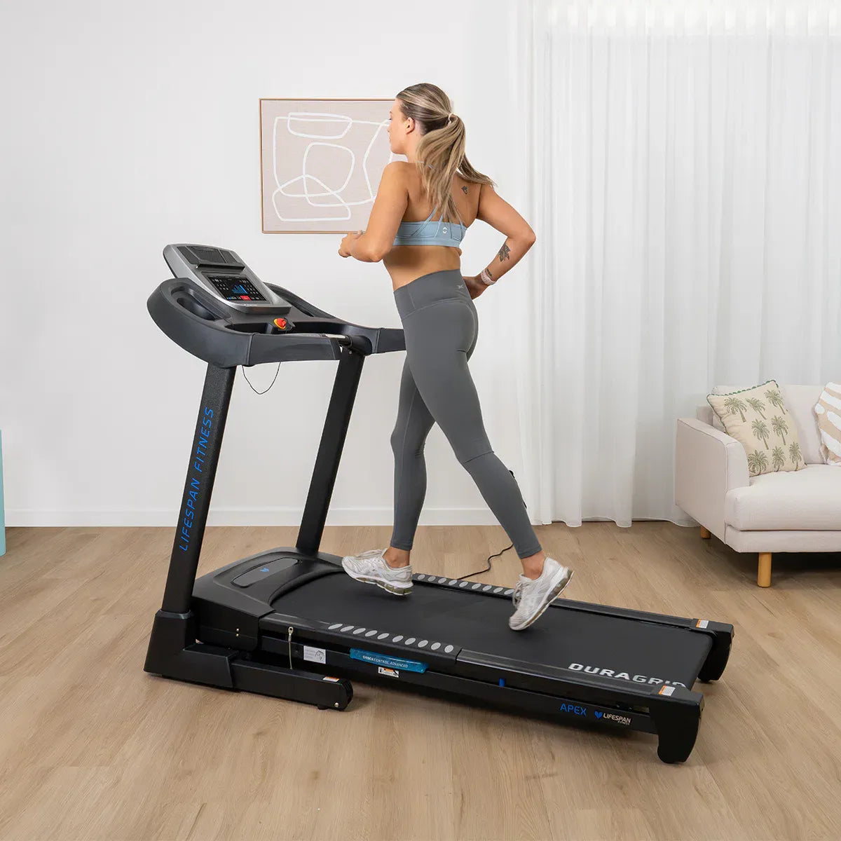 Woman running on Lifespan Apex 2 treadmill in home gym with wooden floor, white curtains and couch