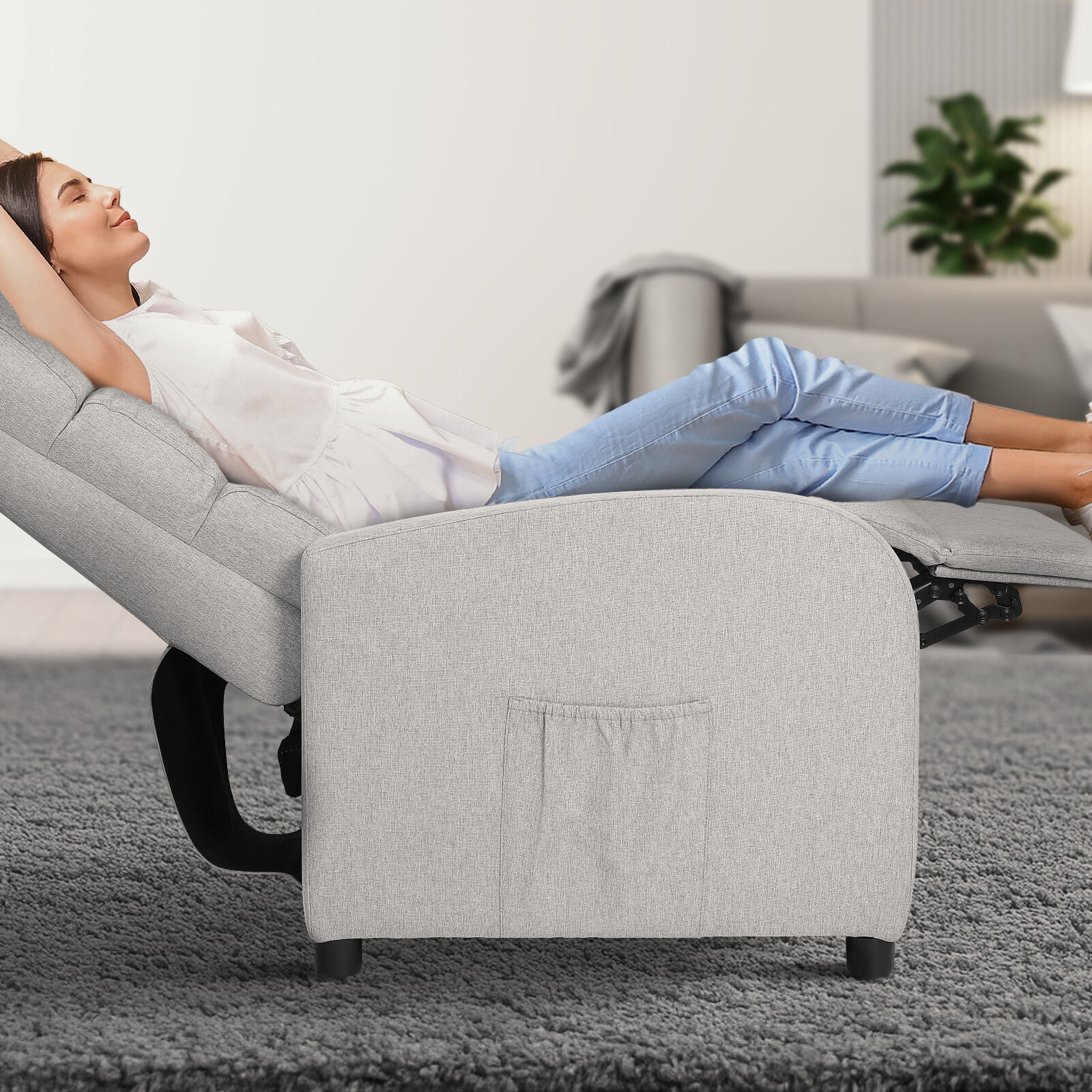 Woman reclining in light grey fabric chair with footrest extended, side pocket visible, living room background