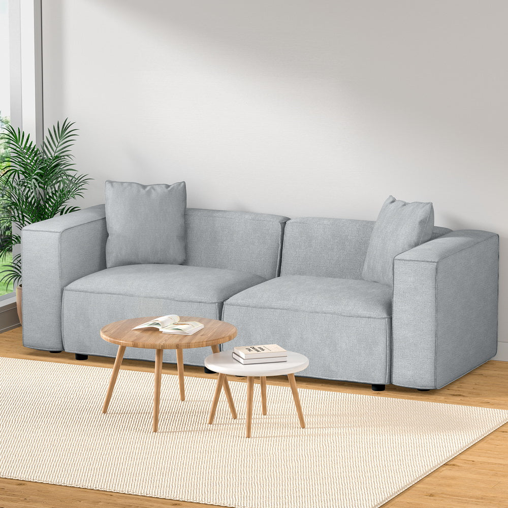 Light grey linen 2-seater modular sofa in bright living room with palm plant and round side tables