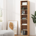 Tall narrow 7-shelf walnut bookcase styled with books and ornaments in bright living room beside armchair and plant