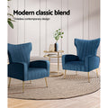 Two navy blue velvet wingback armchairs with gold legs and gold side table on jute rug, 