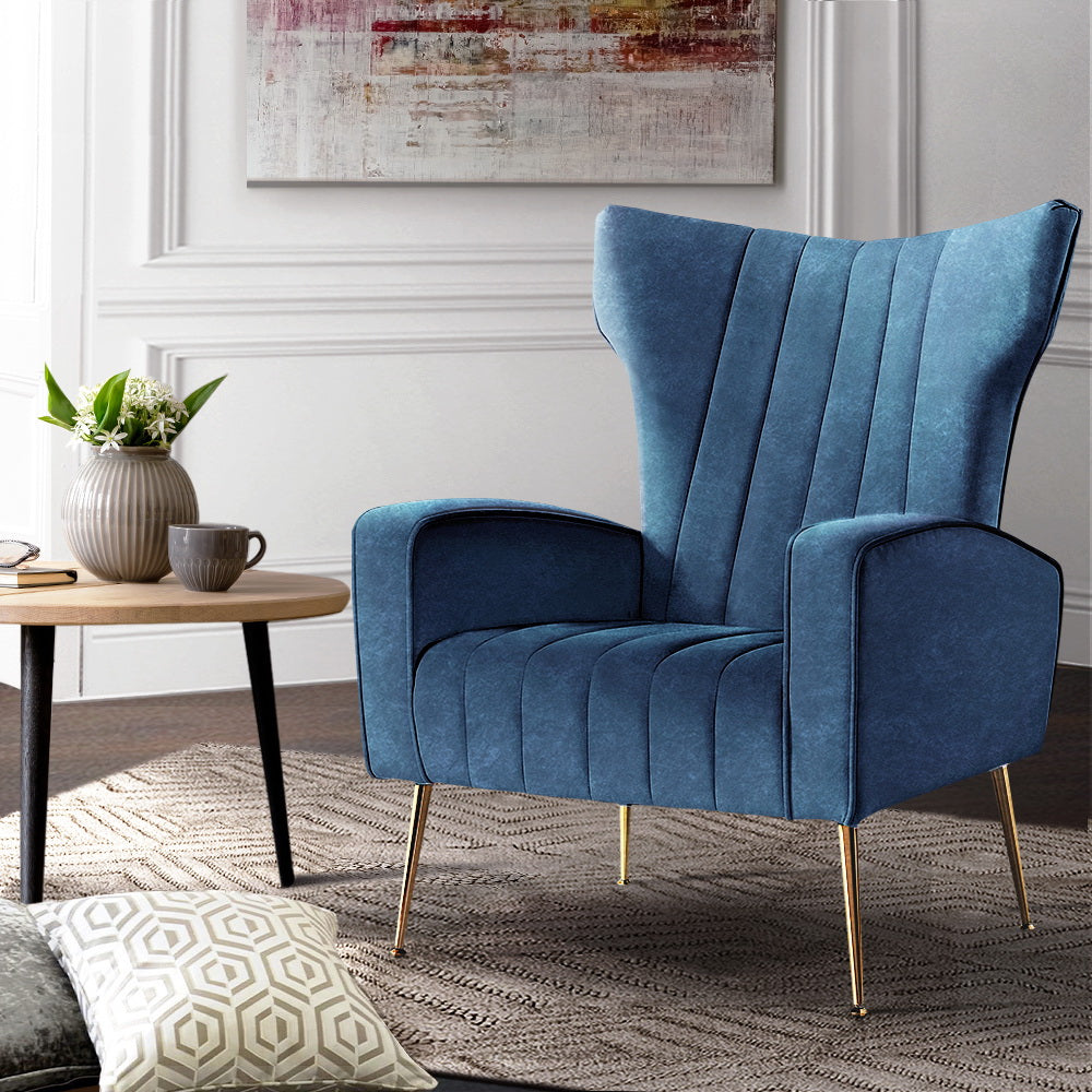 Navy blue velvet wingback armchair with gold legs on textured rug beside round wooden side table, panelled wall