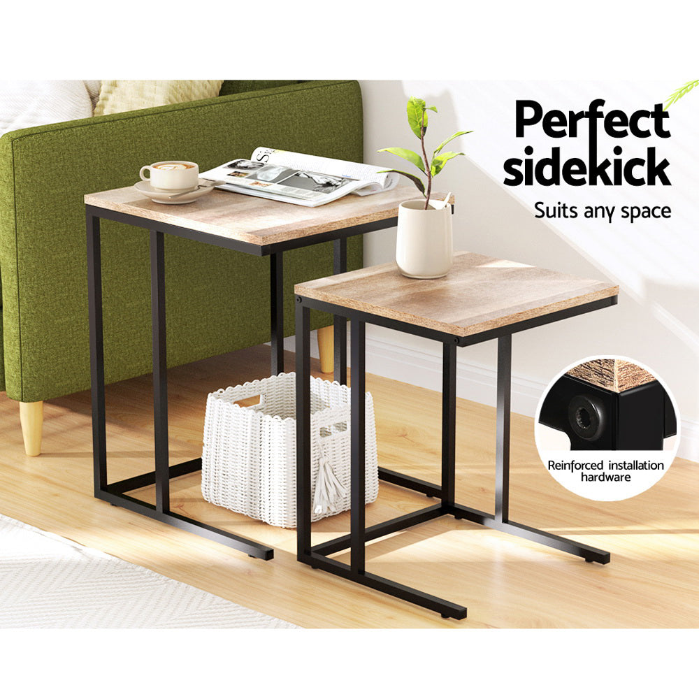 Two nesting C-shaped side tables separated beside a green sofa, one with a coffee cup and magazine, one with a vase.