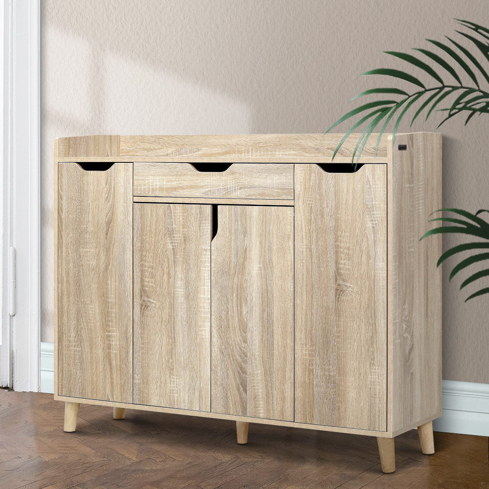 Oak finish shoe cabinet with top drawer and three doors closed in hallway lifestyle with herringbone floor and palm plant