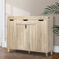 Oak finish shoe cabinet with top drawer and three doors closed in hallway lifestyle with herringbone floor and palm plant