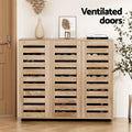 Natural oak shoe cabinet with three slatted ventilated doors loaded with shoes in room with vase and art