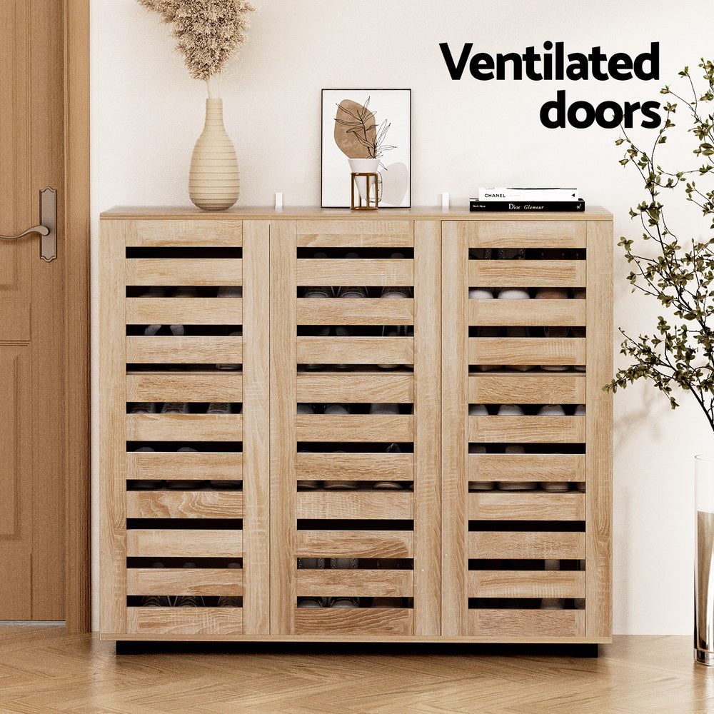 Natural oak shoe cabinet with three slatted ventilated doors loaded with shoes in room with vase and art