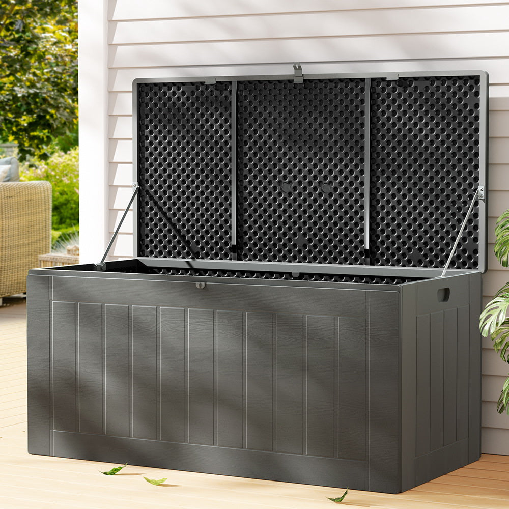 Large grey outdoor storage box with lid open showing ventilated interior, on timber patio with wicker furniture