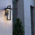 Single black rectangular framed outdoor wall light on rough concrete wall glowing warm, shrub and building visible behind