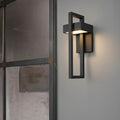 Black rectangular framed outdoor wall light glowing warm on grey wall beside industrial steel-framed glass door panel