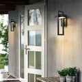 Two black rectangular framed outdoor wall lights flanking a white glass door on covered porch, herb pots on table