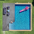Aerial view of backyard pool with dark deck and small wicker sofa set in corner, person on pink inflatable in pool