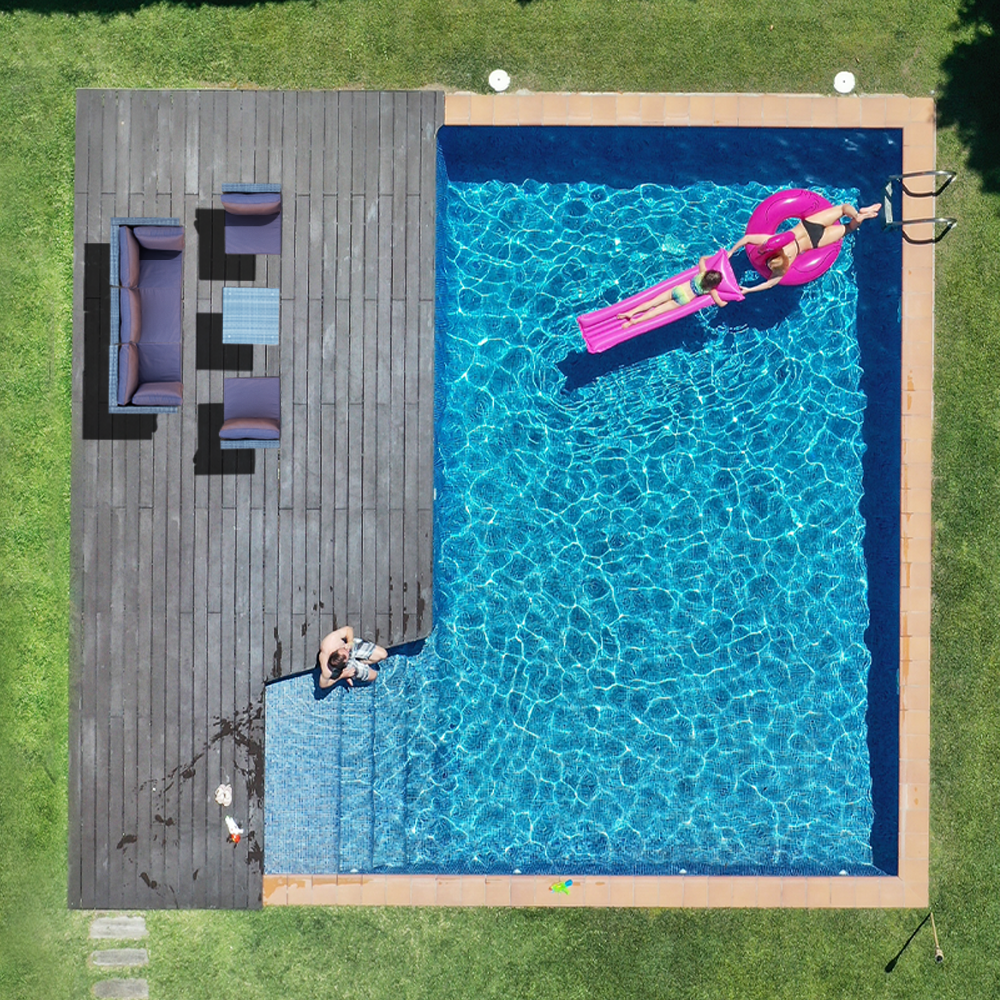 Aerial view of backyard pool with dark deck and small wicker sofa set in corner, person on pink inflatable in pool