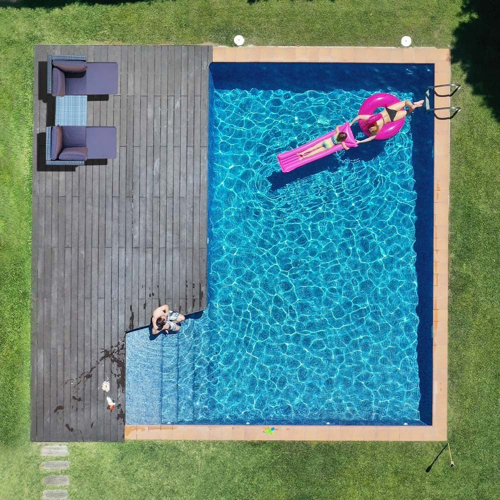 Aerial top-down view of backyard swimming pool with dark deck, dark grey wicker sofa set in corner, person on pink inflatable