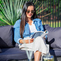 Woman in denim jacket seated on outdoor wicker sofa reading magazine in garden, palm plant visible behind her