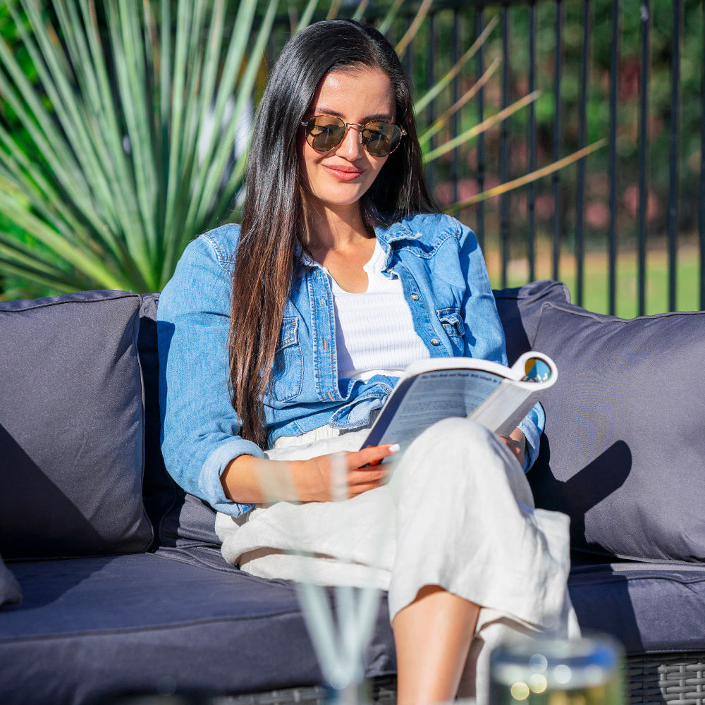 Woman in denim jacket seated on outdoor wicker sofa reading magazine in garden, palm plant visible behind her