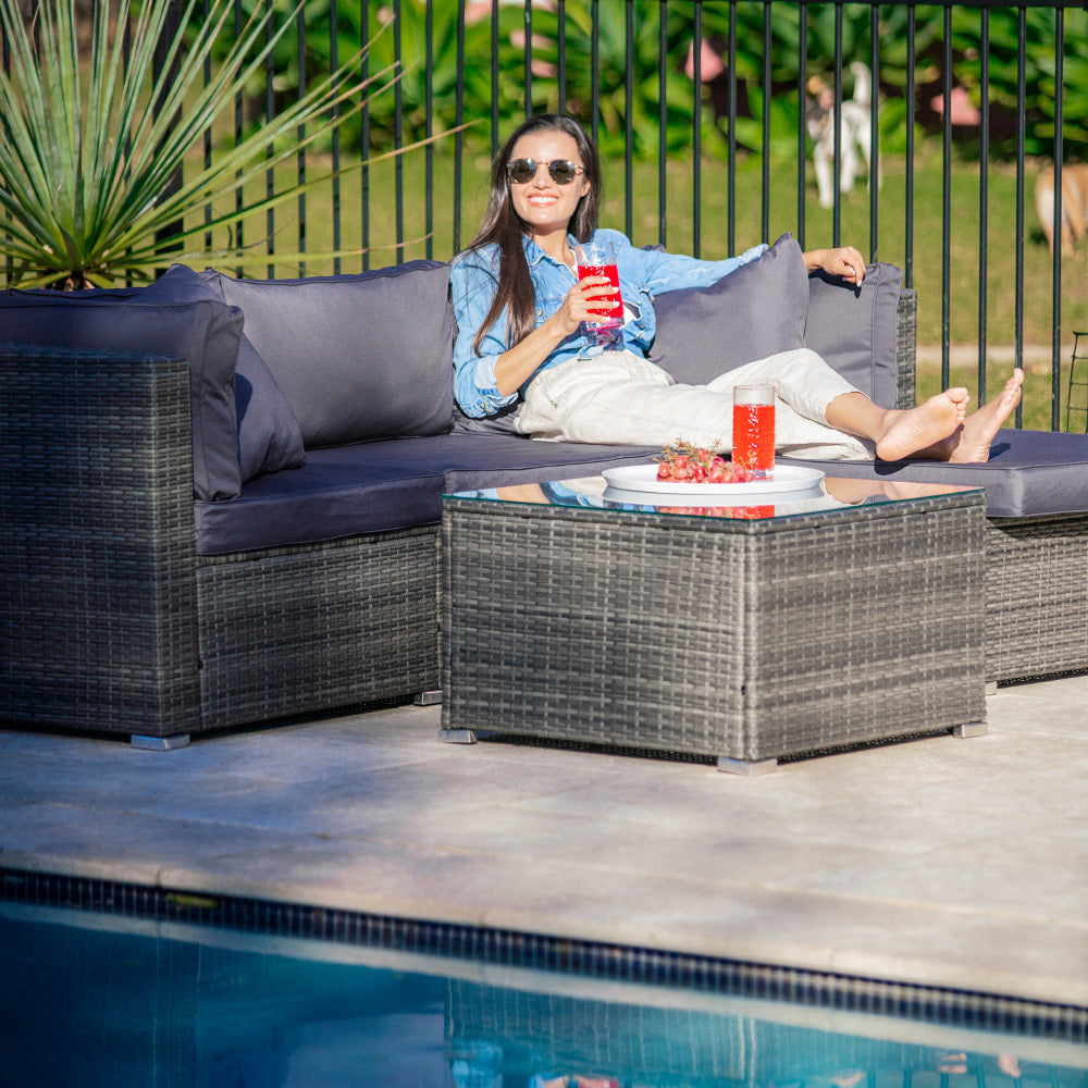 Woman in denim jacket reclining on dark grey wicker outdoor sofa holding red drink beside pool, glass table in foreground