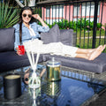 Woman in denim jacket reclining on grey wicker sofa holding red drink, glass coffee table with candle in foreground outdoors
