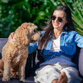 Woman in denim jacket smiling on outdoor wicker sofa with a golden doodle dog beside her, tropical plants in background