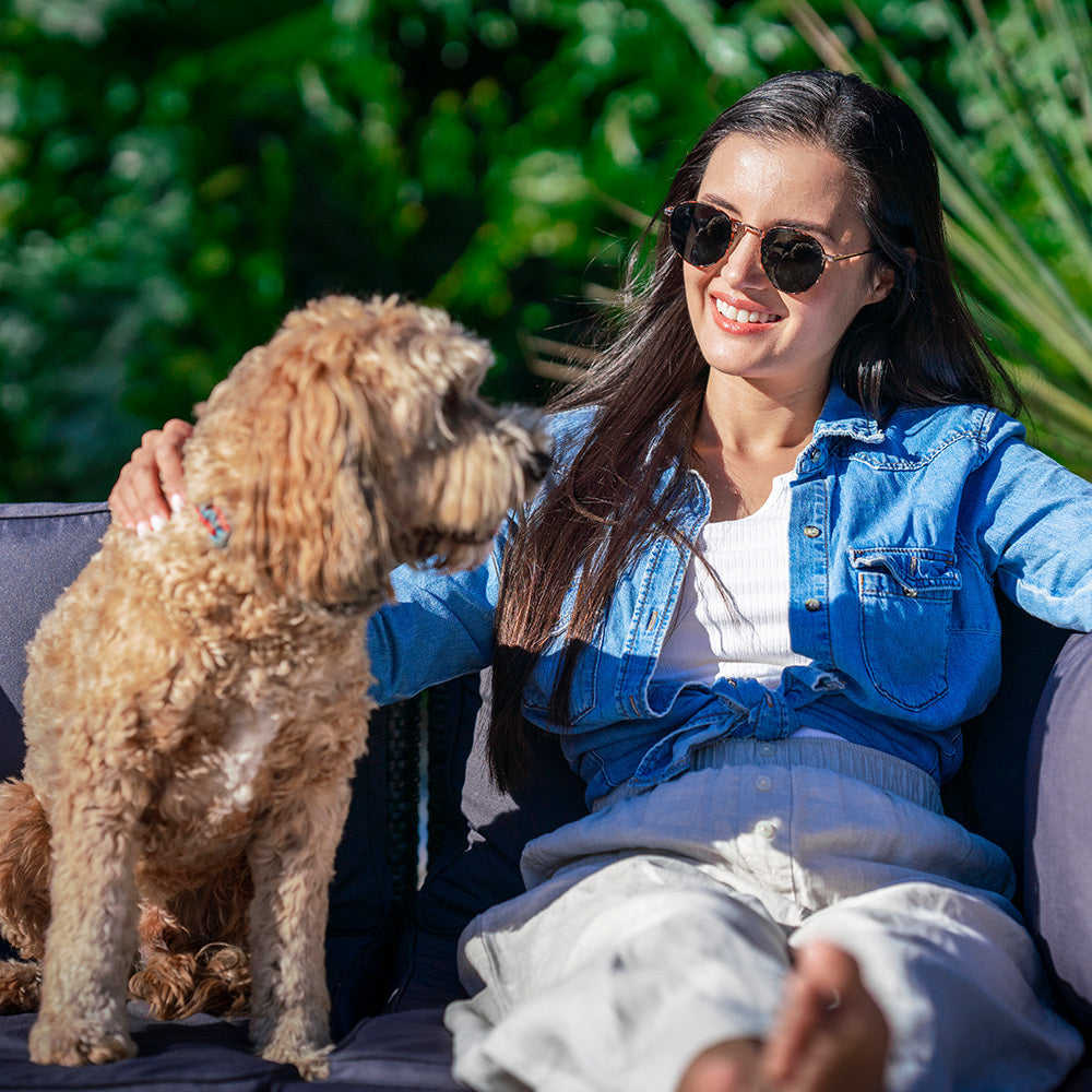 Woman in denim jacket smiling on outdoor wicker sofa with a golden doodle dog beside her, tropical plants in background