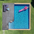 Aerial top-down view of backyard pool with dark deck and wicker sofa set in corner, person on pink pool float and one at edge