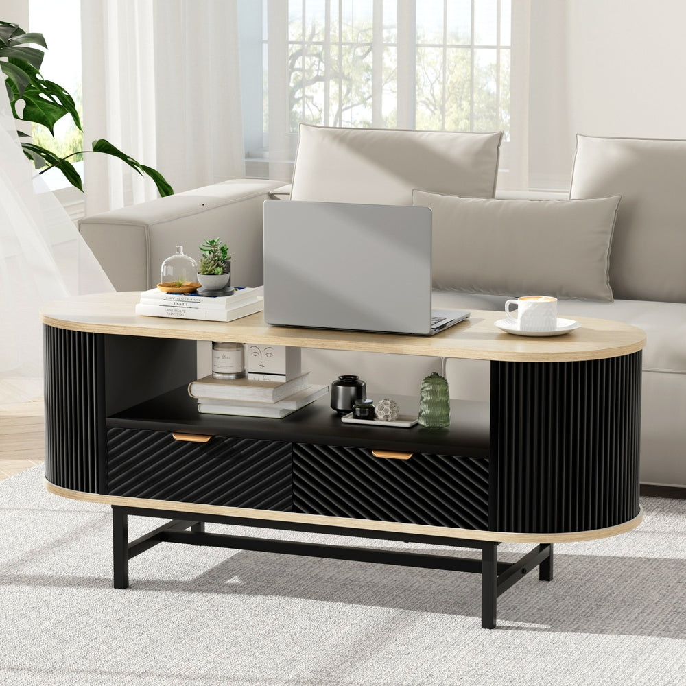 Oval coffee table with black fluted body, oak top, two drawers and open shelf, styled with laptop and plants in a living room