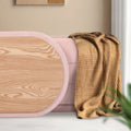 Oval wood grain lid leaning against pink fabric ottoman with tan waffle-knit throw draped over the side
