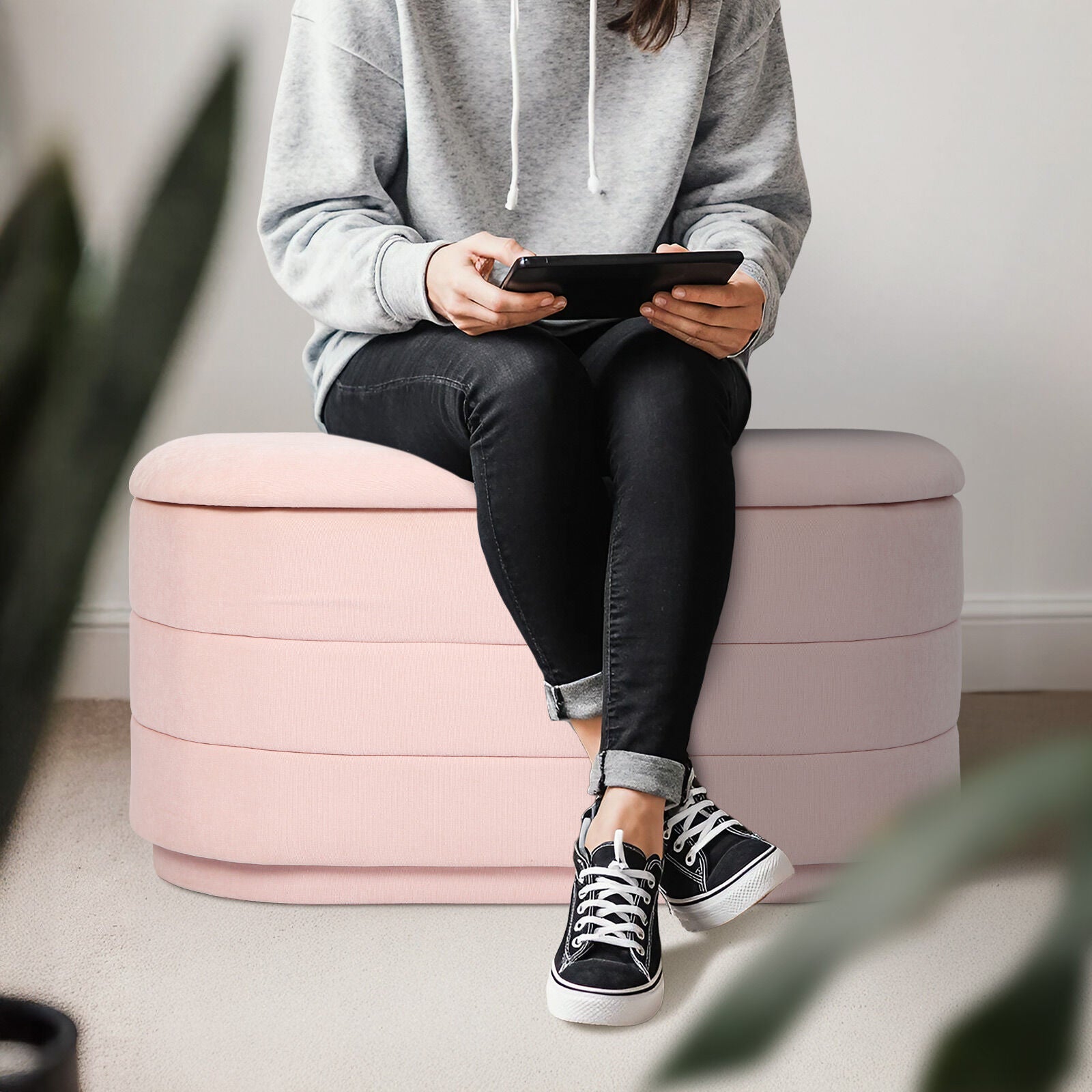 Person in grey hoodie seated on pink fabric storage ottoman holding a tablet, plant visible in foreground