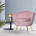 Pink velvet scallop shell armchair with gold legs on grey rug beside black side table with gold lamp against light blue wall