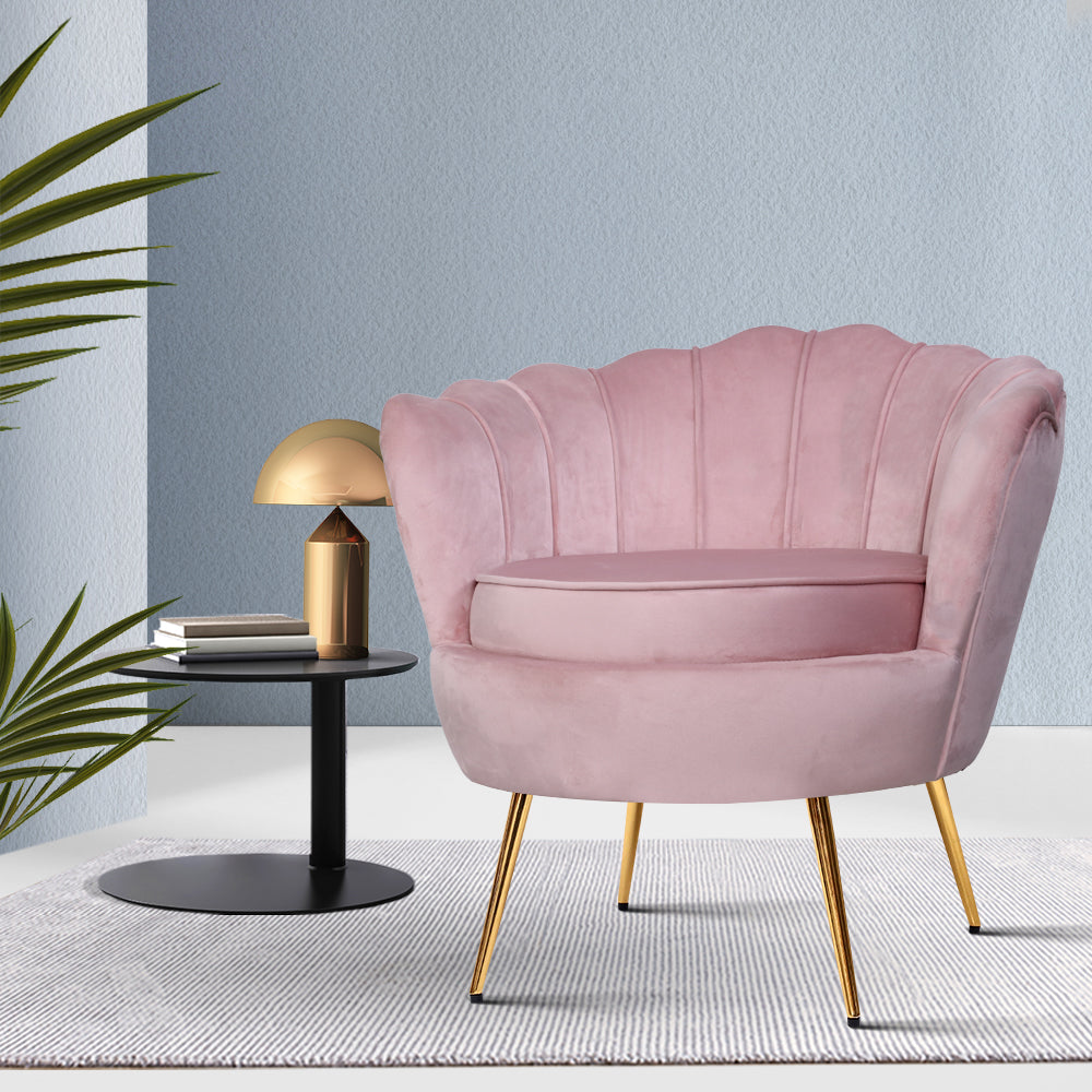Pink velvet scallop shell armchair with gold legs on grey rug beside black side table with gold lamp against light blue wall