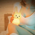 Adult holding warm yellow glowing silicone rabbit night light in bedroom setting, lamp cradled in both hands