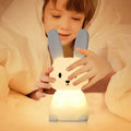 Young boy in bed reaching toward glowing warm white silicone rabbit night light with long blue ears, warm yellow glow
