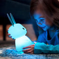 Young girl lying on floor cradling glowing blue silicone rabbit night light, face illuminated in the dark