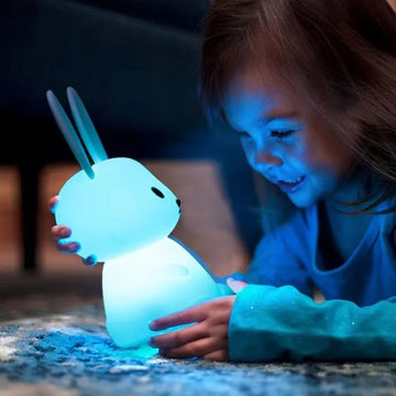 Young girl lying on floor cradling glowing blue silicone rabbit night light, face illuminated in the dark
