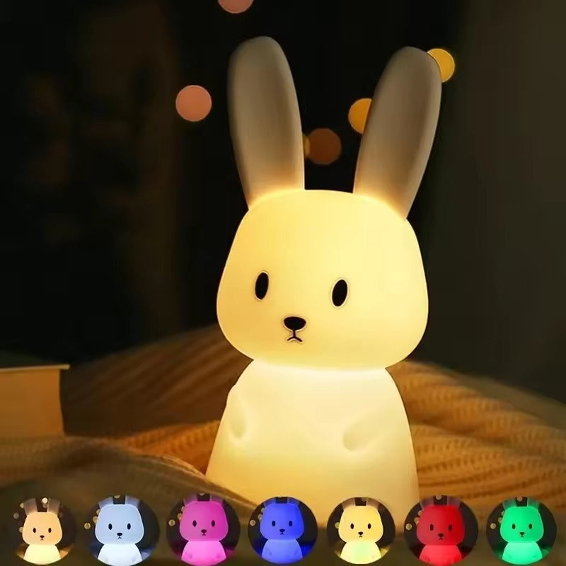 Yellow silicone rabbit night light on knitted surface in dark room, colour mode swatches below: pink, blue, red and green