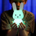 Person cupping hands around glowing teal silicone rabbit night light against blue background, rabbit face visible