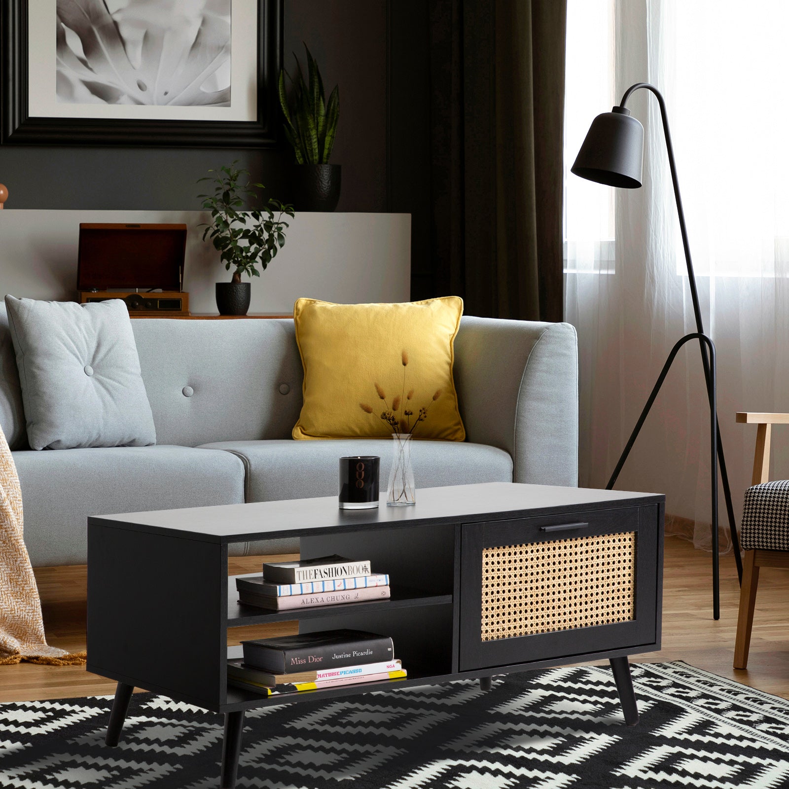 Black rattan coffee table with books on shelves in front of grey sofa with yellow cushion, black floor lamp and geometric rug