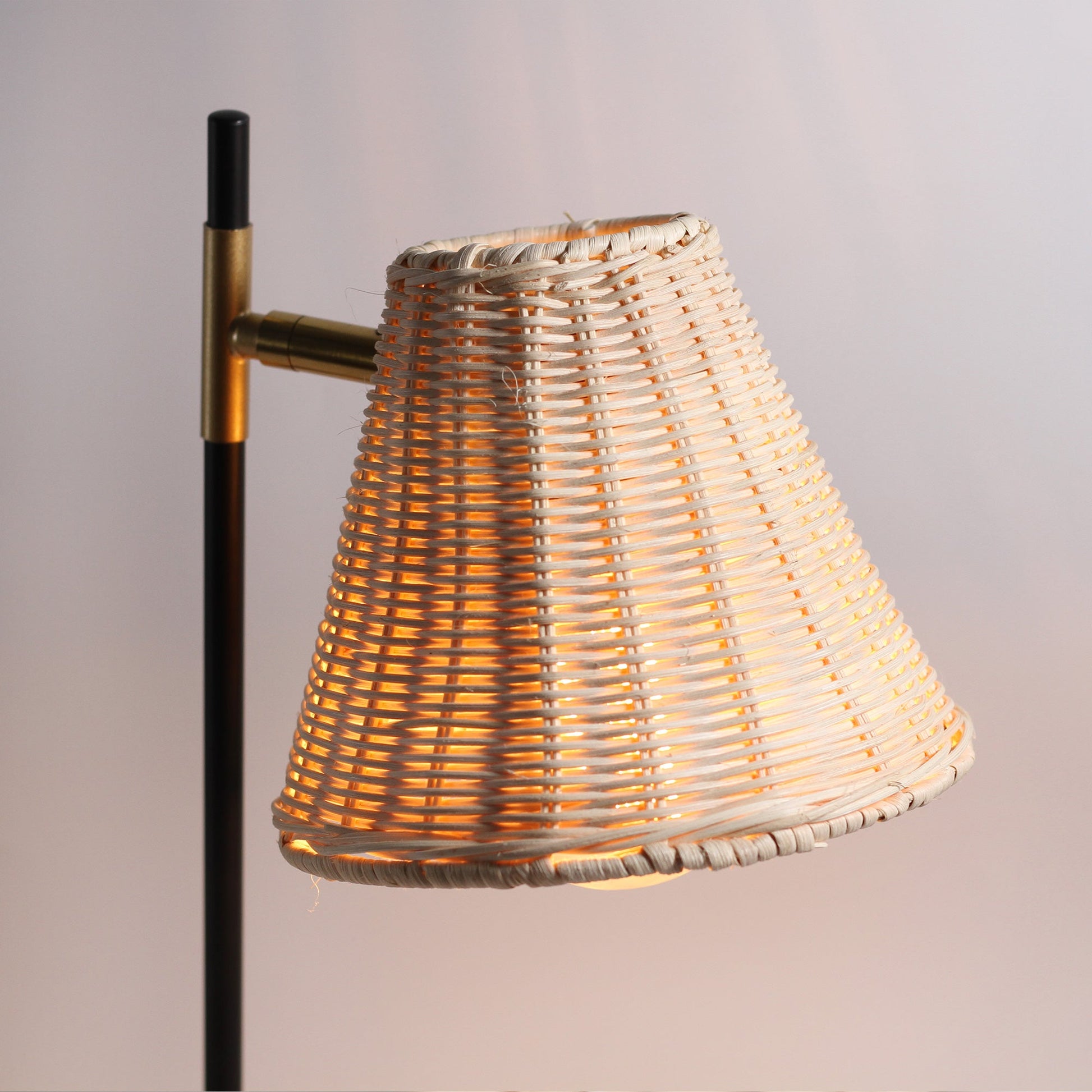 Close-up of woven rattan lampshade glowing warm amber orange light through the weave, brass pivot visible on black pole