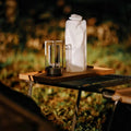 Silver retro lantern LED lamp on wooden camping table beside clear foldable water bottle with outdoor grass background