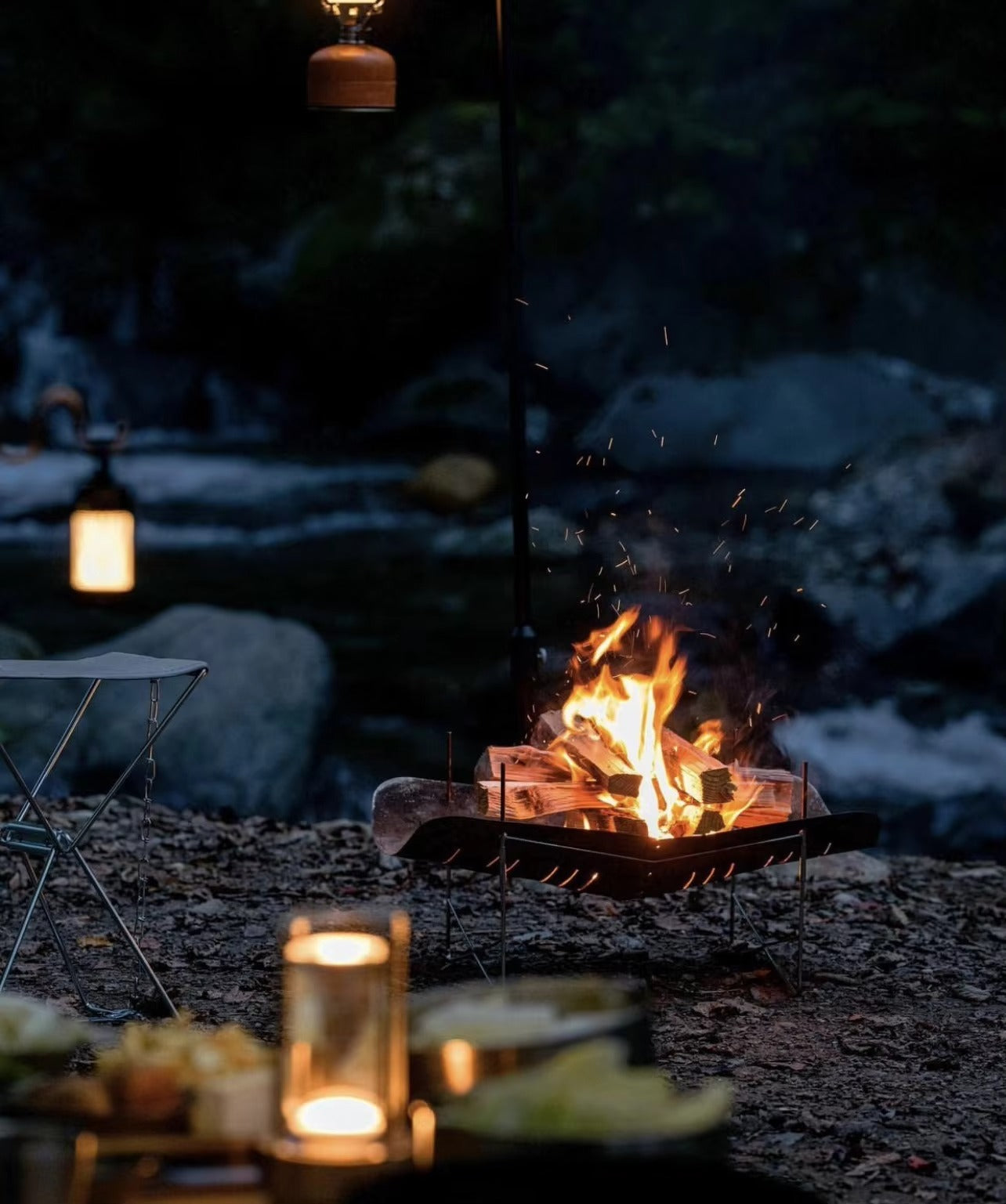 Retro lantern LED lamps glowing at outdoor campsite beside rocky river with campfire in portable grill at night