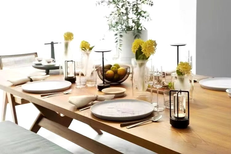 Black retro lantern LED lamp on set wooden dining table with plates, cutlery, glassware and yellow flower vases