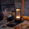Hand holding black retro lantern lamp beside second glowing lamp on wooden board with autumn leaves and oranges