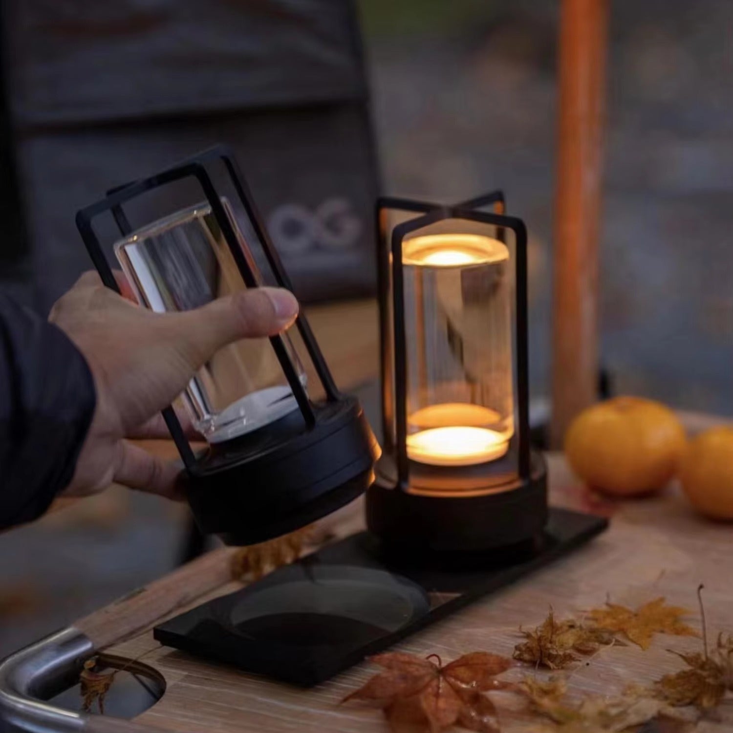 Hand holding black retro lantern lamp beside second glowing lamp on wooden board with autumn leaves and oranges