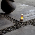 Silver retro lantern LED lamp glowing amber on stone paving with gravel channels and dark ceramic pot in background