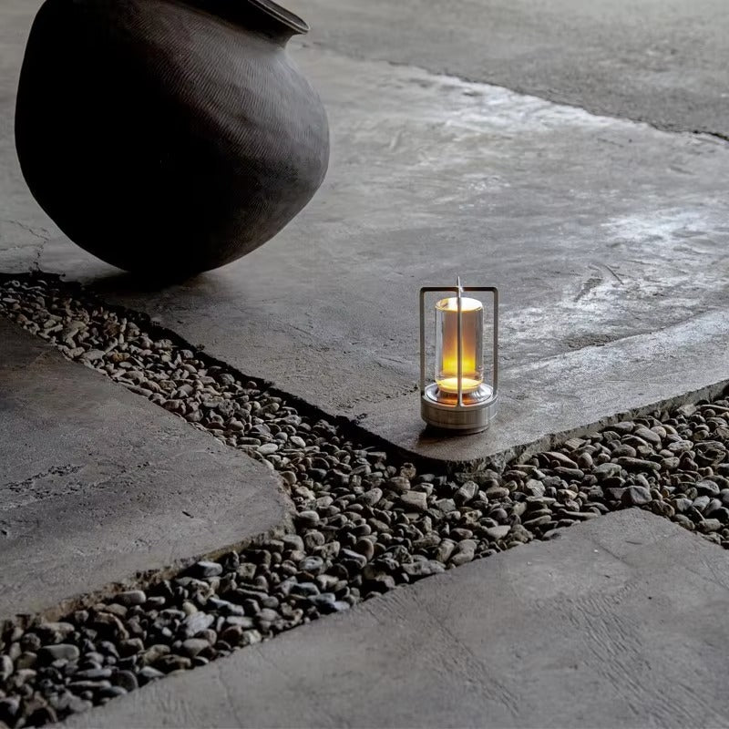 Silver retro lantern LED lamp glowing amber on stone paving with gravel channels and dark ceramic pot in background