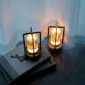 Two black retro lantern LED lamps glowing amber on dark coffee table books against white sheer curtain backdrop
